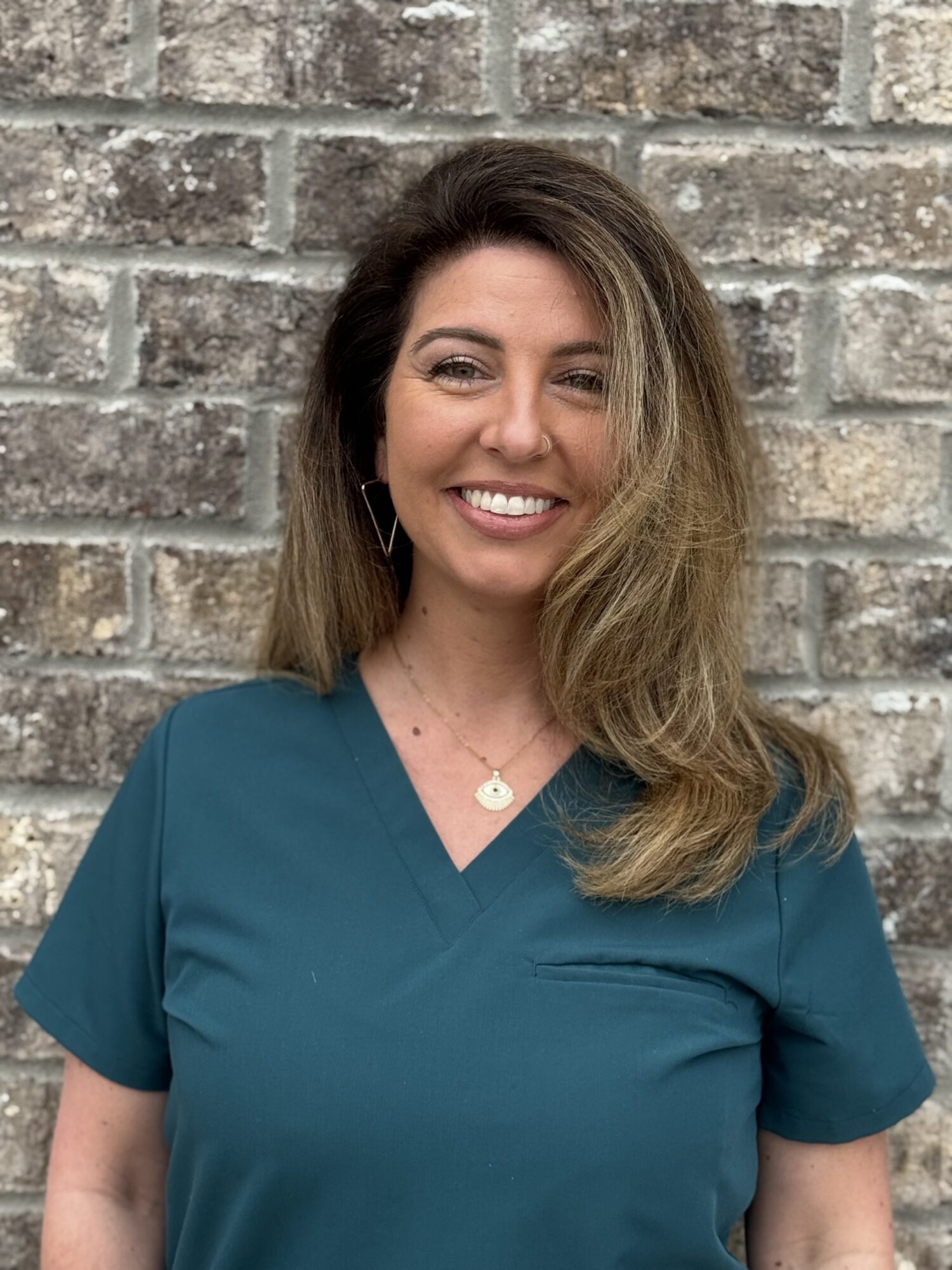 Smiling orthodontic assistant in teal scrubs against a brick wall, representing Shine Pediatric Dentistry & Orthodontics, emphasizing pediatric dental care in Gallatin.