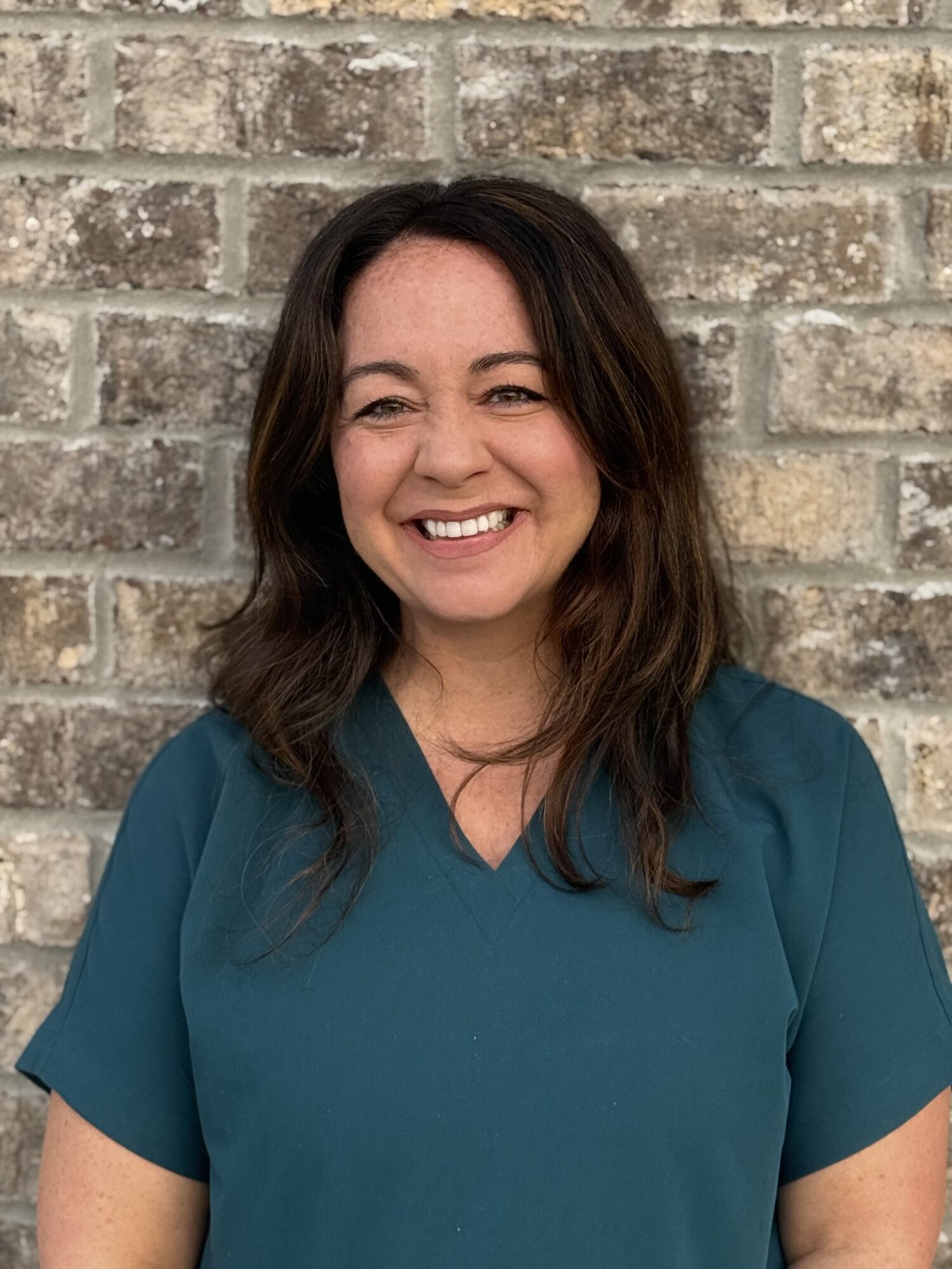 Smiling woman in teal scrubs against a brick wall, representing the friendly staff at Shine Pediatric Dentistry & Orthodontics in Gallatin, TN, focused on pediatric dental care and orthodontics.