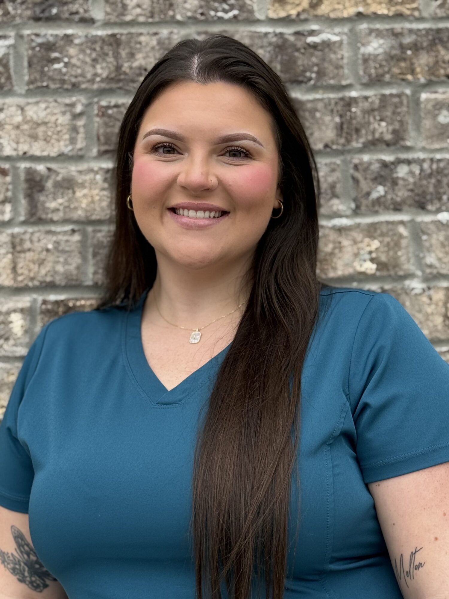 Smiling pediatric dental assistant in teal scrubs against a brick wall, representing Shine Pediatric Dentistry & Orthodontics in Gallatin, focused on children's dental care and orthodontics.