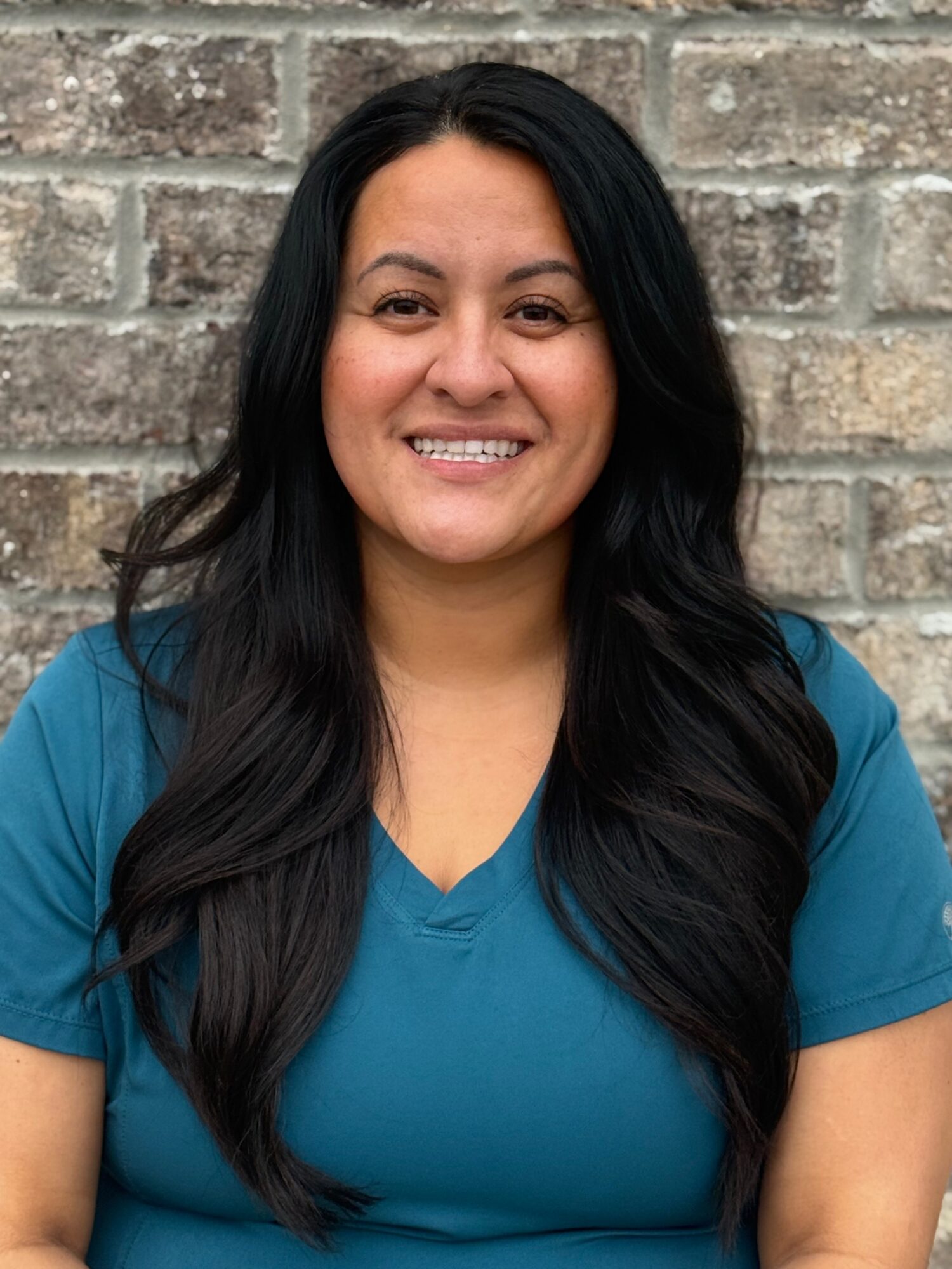 Nicole Vallejos, Orthodontic Treatment Coordinator at Shine Pediatric Dentistry & Orthodontics, smiling in a teal scrub top against a textured brick wall, representing compassionate pediatric dental care in Gallatin, Tennessee.