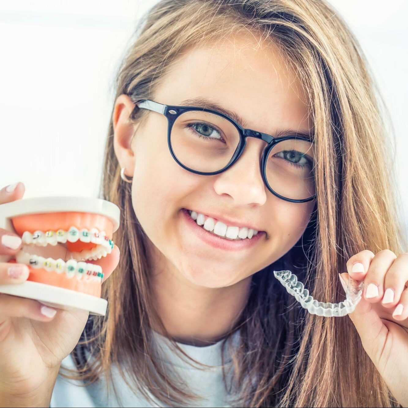 Smiling girl wearing glasses holding a model of braces and a clear aligner, representing orthodontic options at Shine Pediatric Dentistry & Orthodontics in Hendersonville.